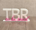 3D printed TBR To Be Read sign in white with hot pink cursive accent, displayed on wooden surface