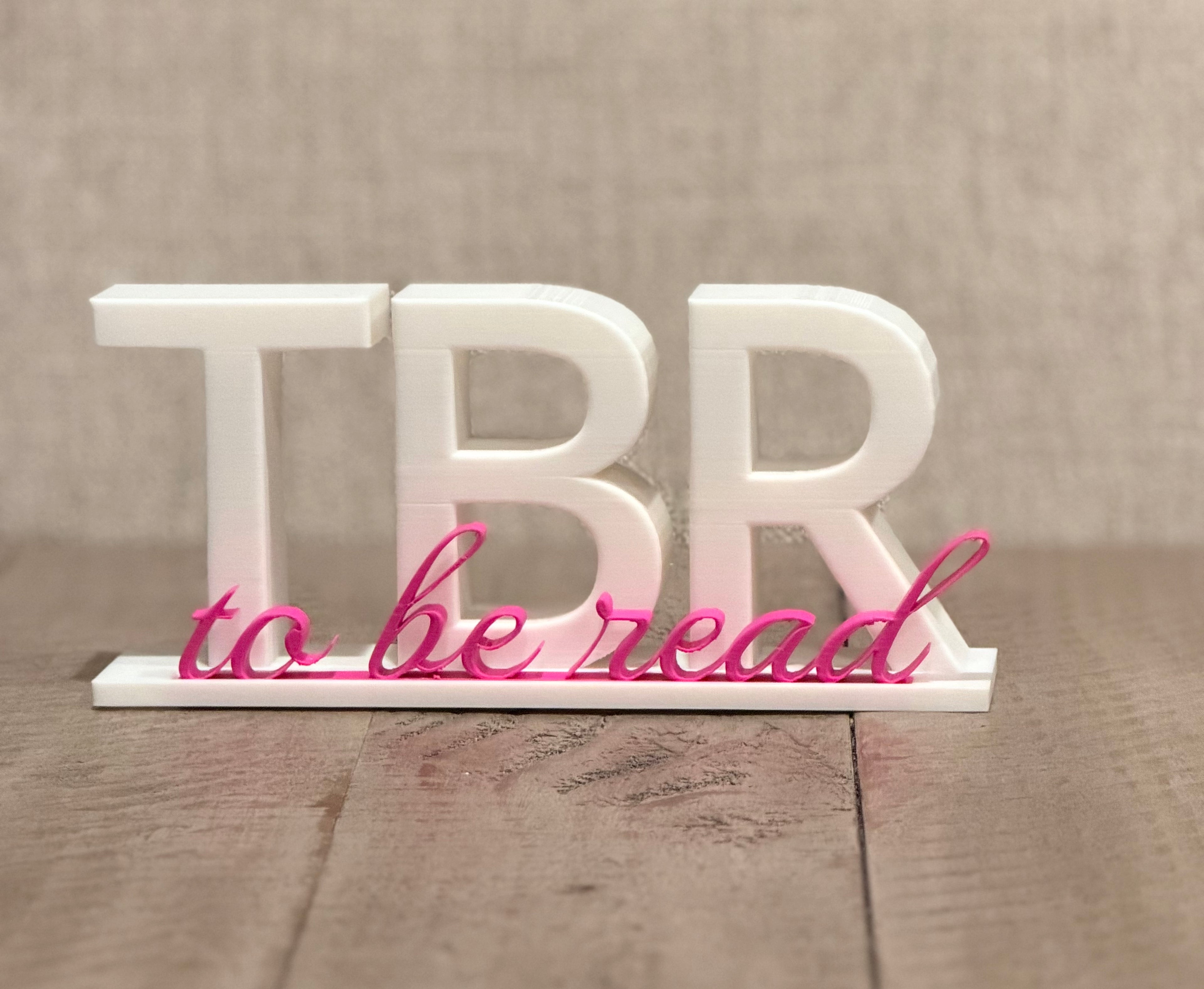 3D printed TBR To Be Read sign in white with hot pink cursive accent, displayed on wooden surface